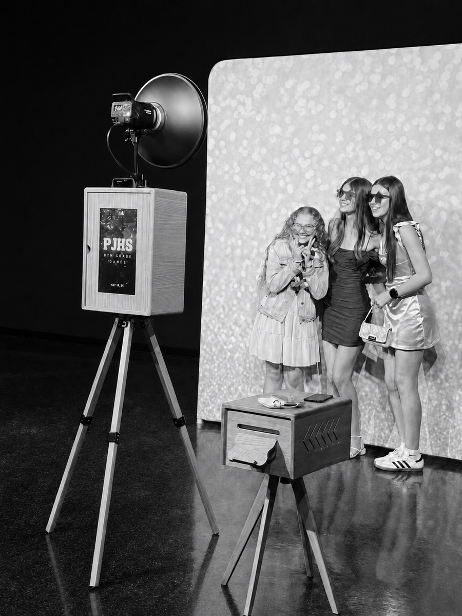 Open-air photo booth with wooden camera, studio lighting, and glitter backdrop at a school dance