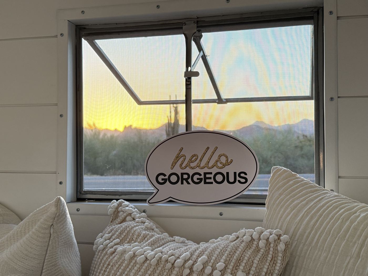 A Hello Gorgeous sign in the trailer window with cozy pillows and a desert sunset